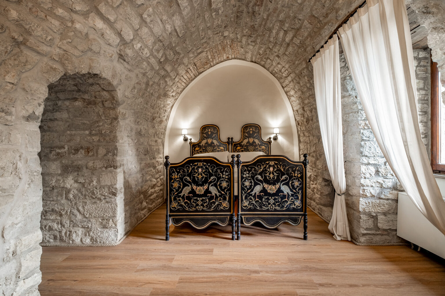 Cozy bedroom featuring vintage black and gold ornate beds, set against a backdrop of rustic stone walls and an arched ceiling. Natural light filters through sheer curtains, enhancing the warm wooden floor and inviting ambiance. Ideal for a charming getaway or historical accommodation experience. | Only on SopranoVillas