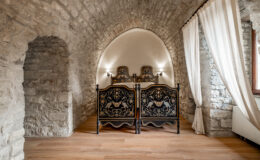 Cozy bedroom featuring vintage black and gold ornate beds, set against a backdrop of rustic stone walls and an arched ceiling. Natural light filters through sheer curtains, enhancing the warm wooden floor and inviting ambiance. Ideal for a charming getaway or historical accommodation experience.