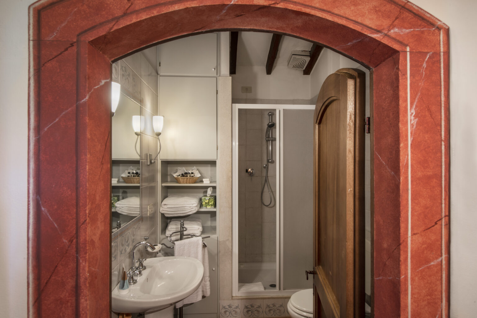 Cozy bathroom interior featuring a red marble archway, a modern shower, a white sink with elegant fixtures, neatly stacked towels, and stylish storage shelves, creating a welcoming and functional space. | Only on SopranoVillas