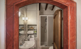 Cozy bathroom interior featuring a red marble archway, a modern shower, a white sink with elegant fixtures, neatly stacked towels, and stylish storage shelves, creating a welcoming and functional space.