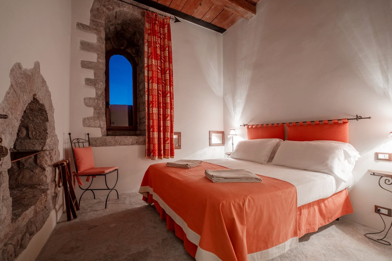 Cozy bedroom with a double bed featuring orange bedding, a decorative headboard, and bedside lamps. The room includes a stone wall with a window, vibrant patterned curtains, and a small chair, creating a warm and inviting atmosphere. Ideal for a relaxing stay. | Only on SopranoVillas