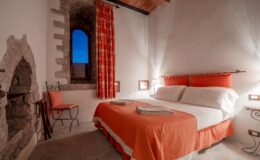Cozy bedroom with a double bed featuring orange bedding, a decorative headboard, and bedside lamps. The room includes a stone wall with a window, vibrant patterned curtains, and a small chair, creating a warm and inviting atmosphere. Ideal for a relaxing stay.