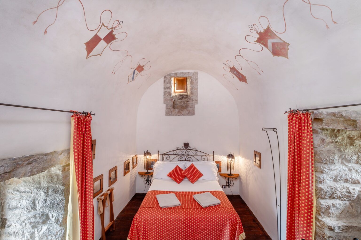 Cozy bedroom with a wrought iron bed adorned with red and white patterned bedding, featuring decorative pillows. The room has arched ceilings with artistic frescoes, stone walls, and warm lighting from lanterns. Red curtains frame the entrance, adding a touch of elegance to the rustic decor. | Only on SopranoVillas