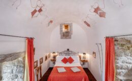 Cozy bedroom with a wrought iron bed adorned with red and white patterned bedding, featuring decorative pillows. The room has arched ceilings with artistic frescoes, stone walls, and warm lighting from lanterns. Red curtains frame the entrance, adding a touch of elegance to the rustic decor.