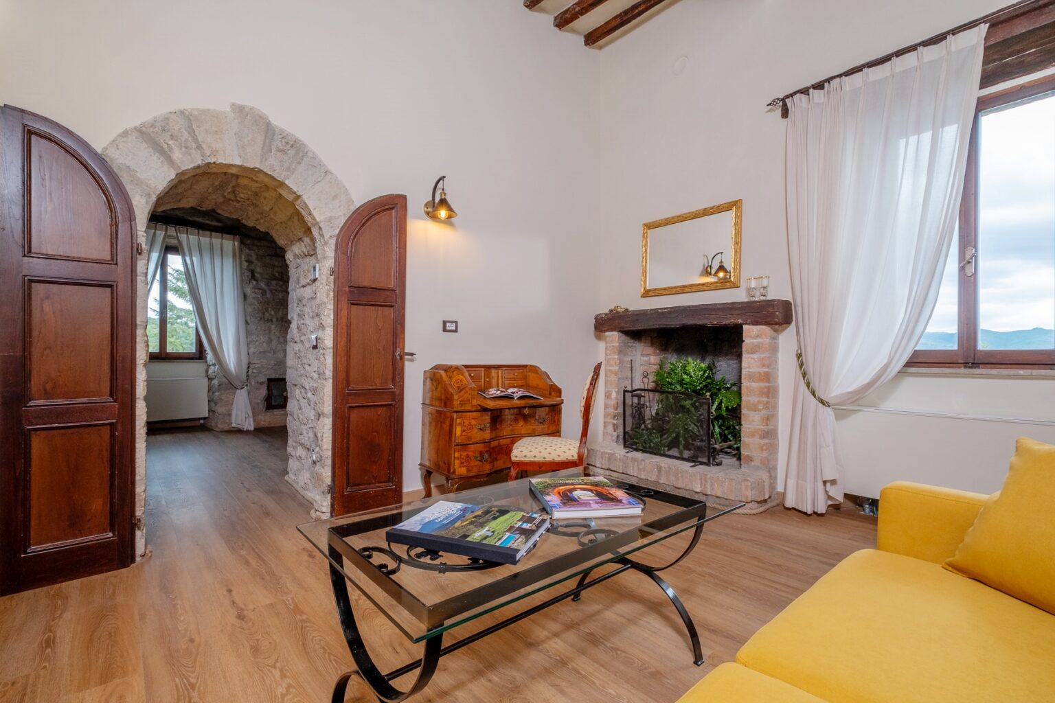 Cozy living room featuring rustic stone archways, wooden doors, and a warm fireplace with decorative plants. A glass coffee table holds travel magazines, and a comfortable yellow sofa invites relaxation. Natural light streams through the large window, enhancing the inviting atmosphere. | Only on SopranoVillas