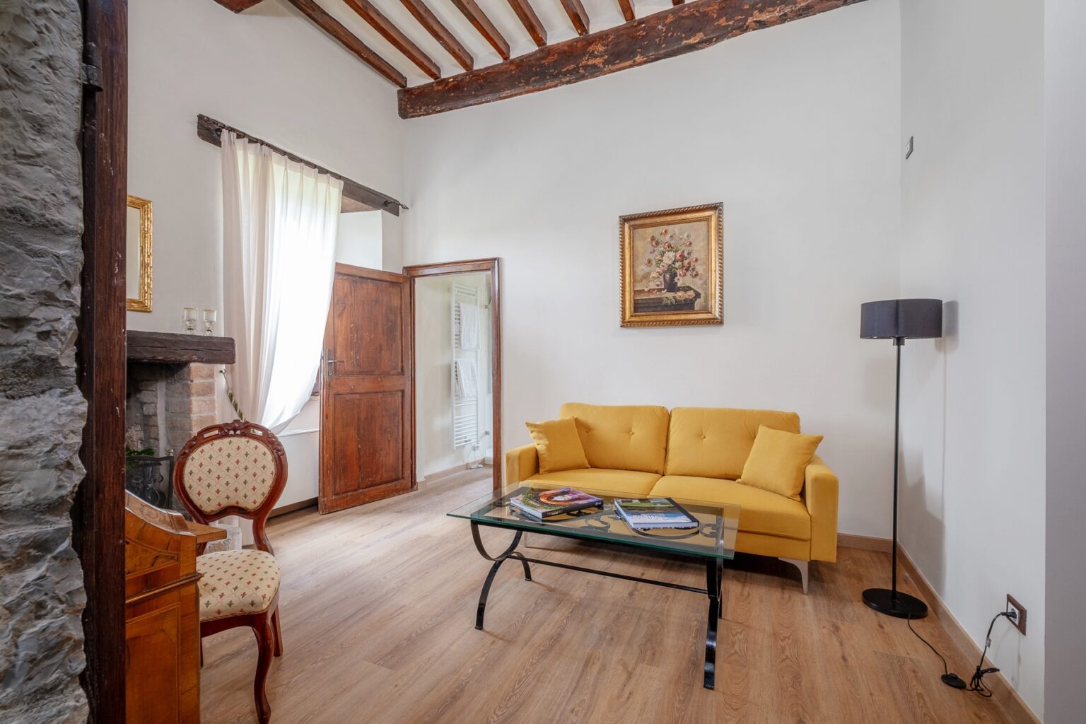 Cozy living room featuring a bright yellow sofa, a glass coffee table with magazines, and a vintage wooden chair. Natural light filters through sheer curtains, highlighting the rustic wooden beams and stone wall. A framed floral painting adds an artistic touch to the space. | Only on SopranoVillas