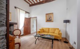 Cozy living room featuring a bright yellow sofa, a glass coffee table with magazines, and a vintage wooden chair. Natural light filters through sheer curtains, highlighting the rustic wooden beams and stone wall. A framed floral painting adds an artistic touch to the space.