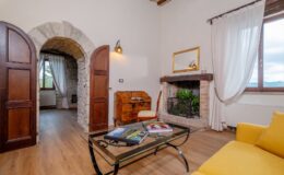 Cozy living room featuring rustic stone archways, wooden doors, and a warm fireplace with decorative plants. A glass coffee table holds travel magazines, and a comfortable yellow sofa invites relaxation. Natural light streams through the large window, enhancing the inviting atmosphere.