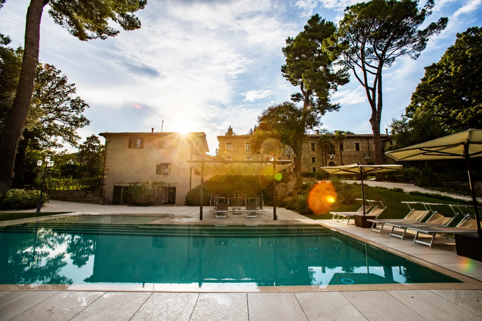 Scenic view of a luxurious swimming pool surrounded by lush greenery and elegant sun loungers, with a charming villa in the background, capturing a beautiful sunset ambiance. | Only on SopranoVillas