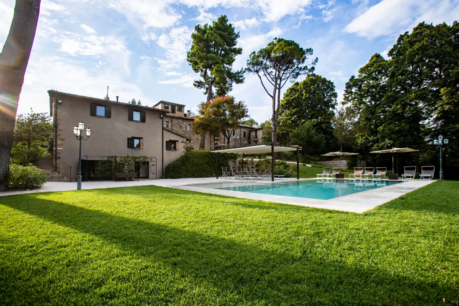 Scenic view of a modern villa with a swimming pool surrounded by lush green grass and tall trees, featuring lounge chairs and umbrellas for relaxation. The house showcases a blend of natural stone and contemporary design elements, set against a clear blue sky. | Only on SopranoVillas
