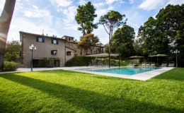 Scenic view of a modern villa with a swimming pool surrounded by lush green grass and tall trees, featuring lounge chairs and umbrellas for relaxation. The house showcases a blend of natural stone and contemporary design elements, set against a clear blue sky.