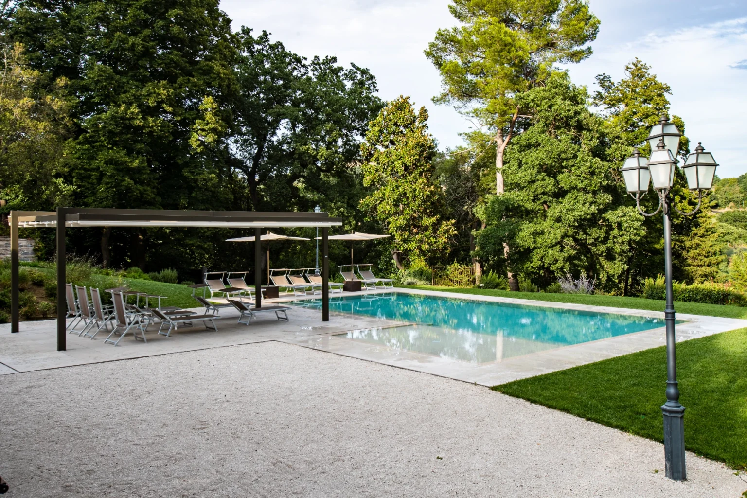 Modern outdoor swimming pool surrounded by lush greenery, featuring lounge chairs, umbrellas, and a stylish pergola for shade. Elegant lamp post adds to the serene atmosphere. Ideal for relaxation and leisure. | Only on SopranoVillas