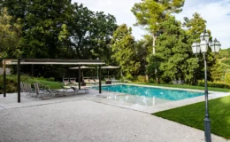 Modern outdoor swimming pool surrounded by lush greenery, featuring lounge chairs, umbrellas, and a stylish pergola for shade. Elegant lamp post adds to the serene atmosphere. Ideal for relaxation and leisure.