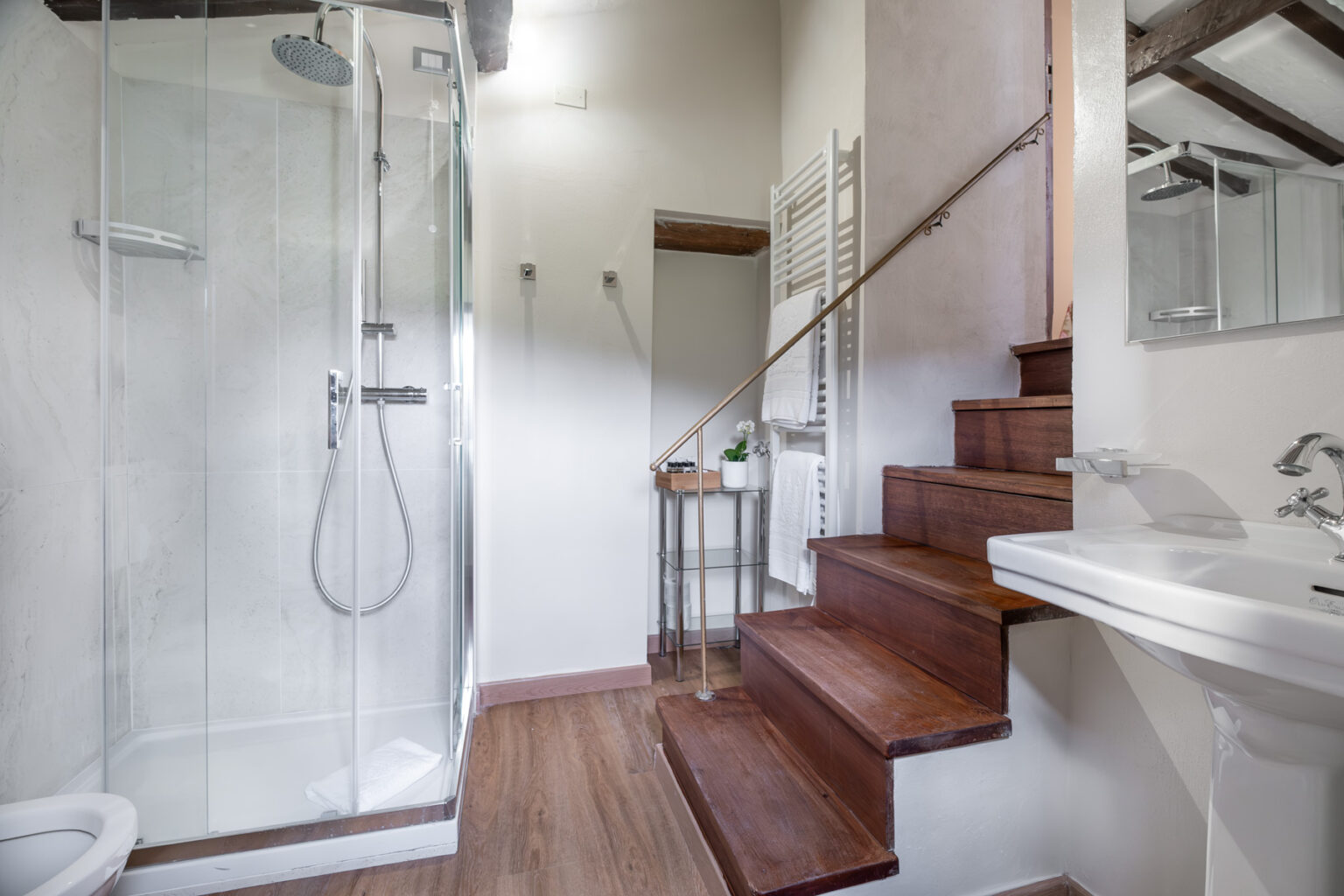 Modern bathroom featuring a glass shower enclosure, wooden staircase, and stylish sink. The space includes a heated towel rack and a small decorative plant, creating a warm and inviting atmosphere. | Only on SopranoVillas