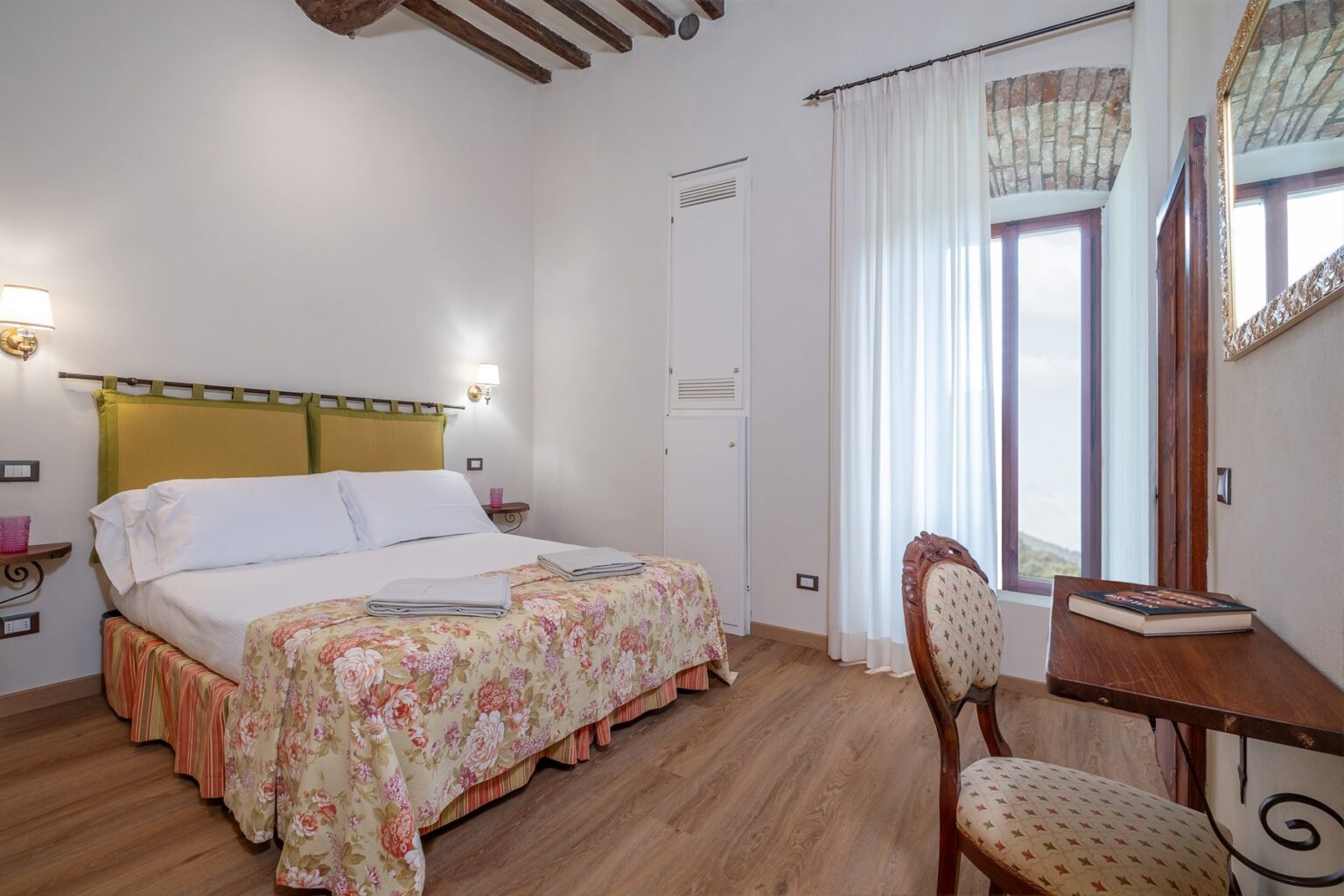 Cozy hotel room featuring a double bed with floral bedding, wooden beams, and large windows with sheer curtains. A small wooden desk and chair are positioned near the window, creating a comfortable and inviting atmosphere for relaxation. | Only on SopranoVillas