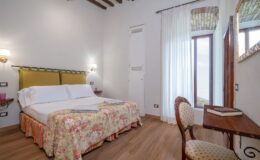 Cozy hotel room featuring a double bed with floral bedding, wooden beams, and large windows with sheer curtains. A small wooden desk and chair are positioned near the window, creating a comfortable and inviting atmosphere for relaxation.
