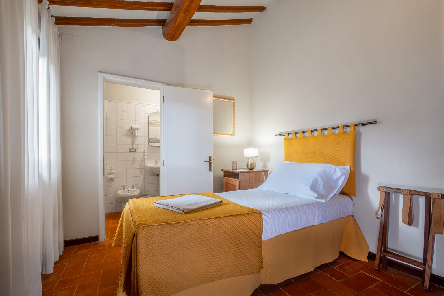 Cozy bedroom featuring a single bed with a yellow bedspread, wooden beams on the ceiling, and natural light from sheer curtains. A small wooden nightstand with a lamp is beside the bed, and a bathroom is visible through an open door. Ideal for a comfortable stay. | Only on SopranoVillas