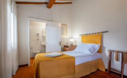 Cozy bedroom featuring a single bed with a yellow bedspread, wooden beams on the ceiling, and natural light from sheer curtains. A small wooden nightstand with a lamp is beside the bed, and a bathroom is visible through an open door. Ideal for a comfortable stay.