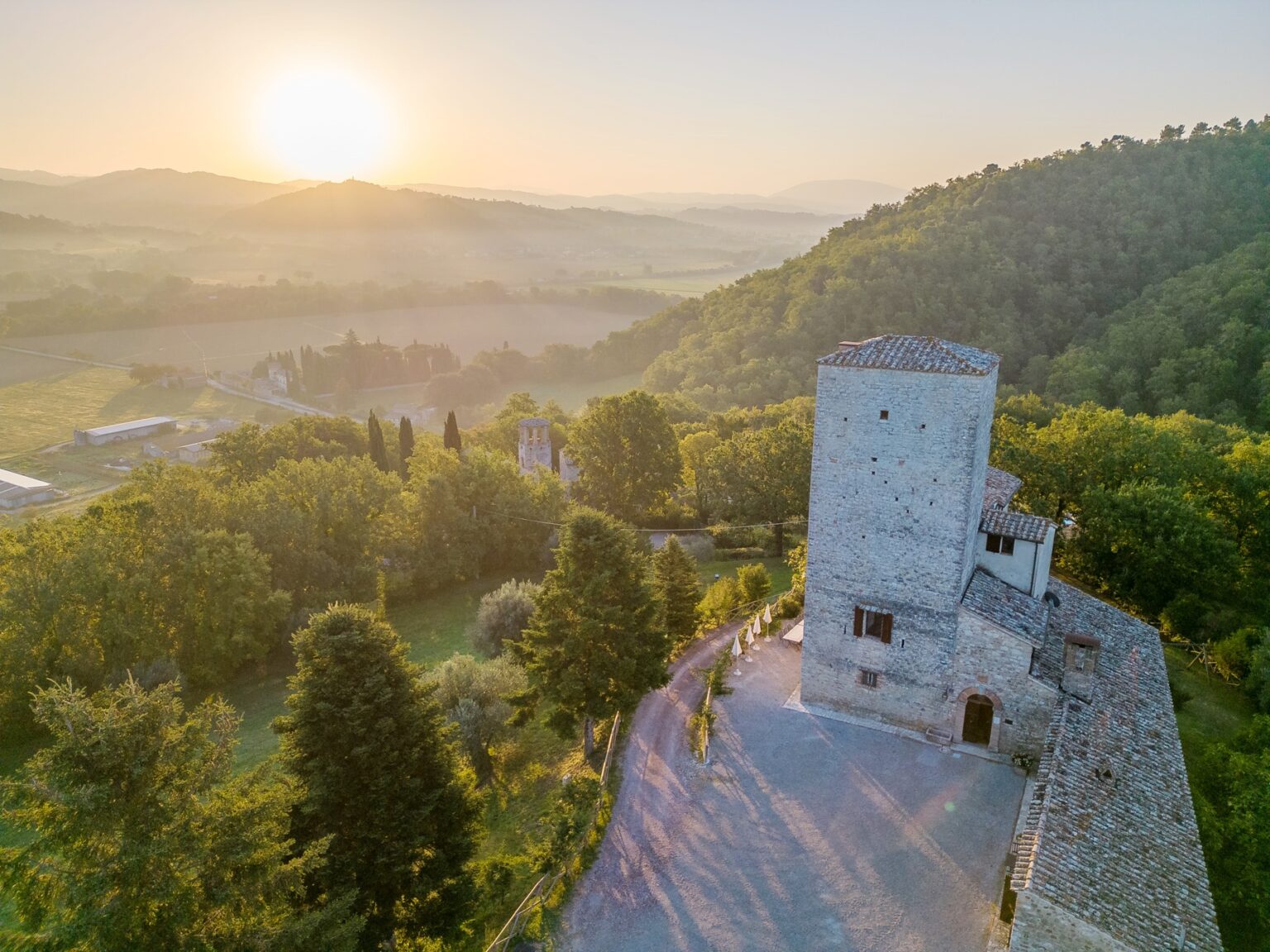 Aerial view of a historic stone tower and surrounding landscape at sunrise, featuring lush green trees and rolling hills in the background, capturing the serene beauty of the Italian countryside. | Only on SopranoVillas
