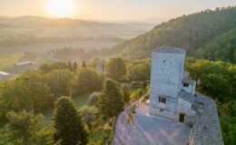 Aerial view of a historic stone tower and surrounding landscape at sunrise, featuring lush green trees and rolling hills in the background, capturing the serene beauty of the Italian countryside.