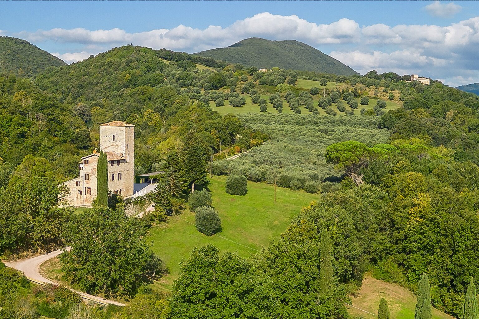 Scenic view of a historic stone tower house surrounded by lush greenery and rolling hills, featuring olive groves and trees under a partly cloudy sky. Ideal for showcasing rural architecture and natural landscapes. | Only on SopranoVillas