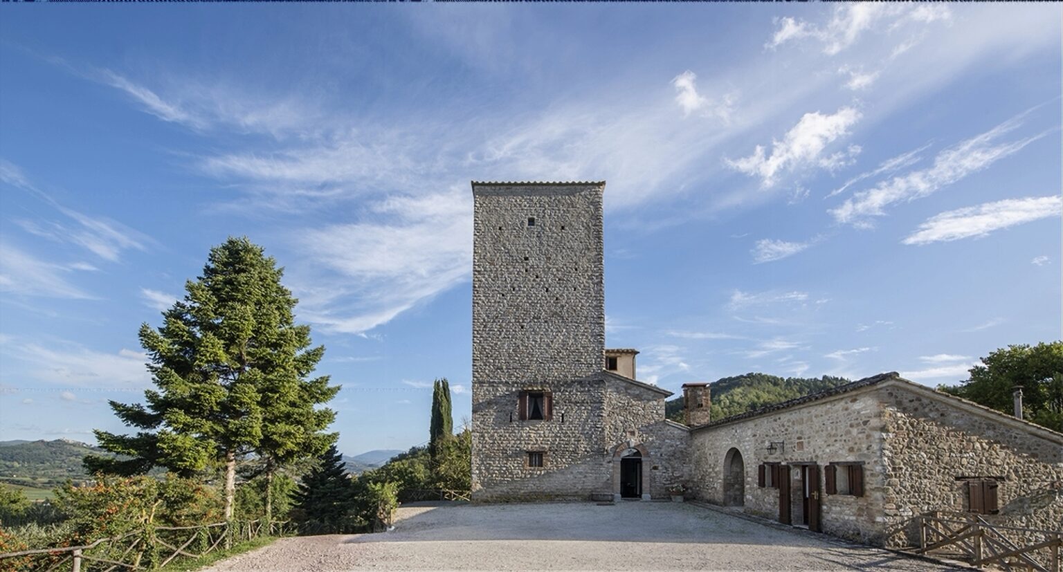 Historic stone tower and building set against a blue sky, surrounded by lush greenery, showcasing traditional architecture in a scenic landscape. Ideal for exploring Italian heritage and countryside. | Only on SopranoVillas