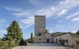 Historic stone tower and building set against a blue sky, surrounded by lush greenery, showcasing traditional architecture in a scenic landscape. Ideal for exploring Italian heritage and countryside.