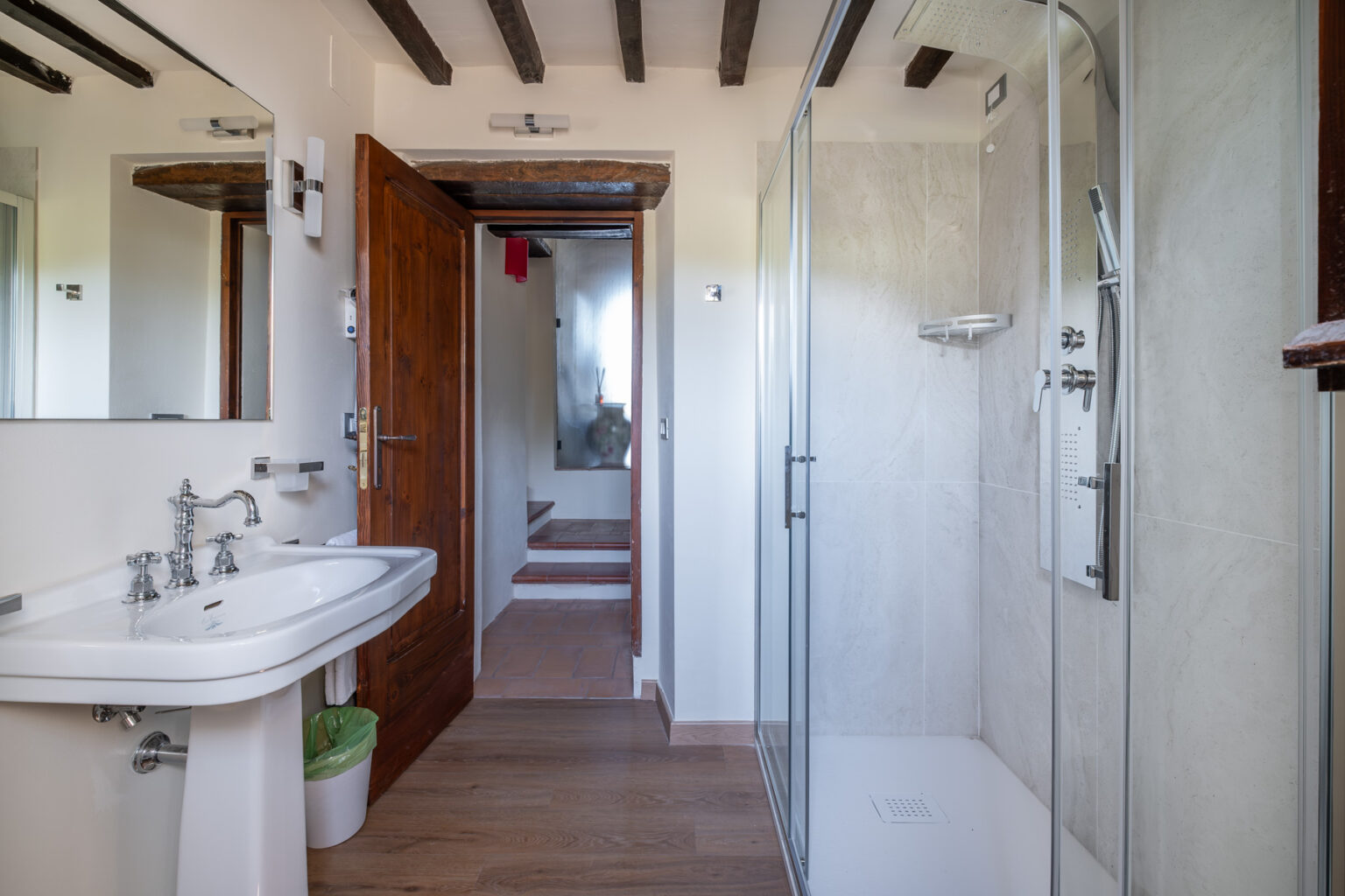 Modern bathroom interior featuring a stylish sink with vintage faucets, a glass shower enclosure, wooden beams, and natural light. The design combines contemporary and rustic elements, showcasing a clean and inviting space. | Only on SopranoVillas