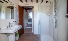 Modern bathroom interior featuring a stylish sink with vintage faucets, a glass shower enclosure, wooden beams, and natural light. The design combines contemporary and rustic elements, showcasing a clean and inviting space.