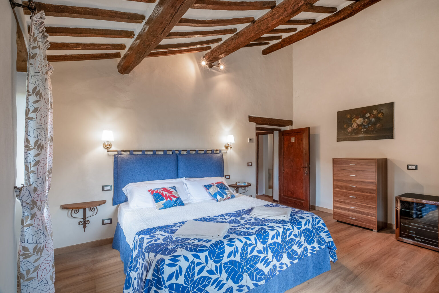 Cozy bedroom featuring a blue upholstered bed with decorative pillows, wooden beams on the ceiling, and a warm color palette. The room includes a wooden dresser, bedside lamps, and a window with patterned curtains, creating a welcoming atmosphere. Ideal for relaxation and comfort. | Only on SopranoVillas