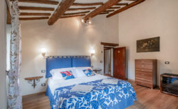 Cozy bedroom featuring a blue upholstered bed with decorative pillows, wooden beams on the ceiling, and a warm color palette. The room includes a wooden dresser, bedside lamps, and a window with patterned curtains, creating a welcoming atmosphere. Ideal for relaxation and comfort.