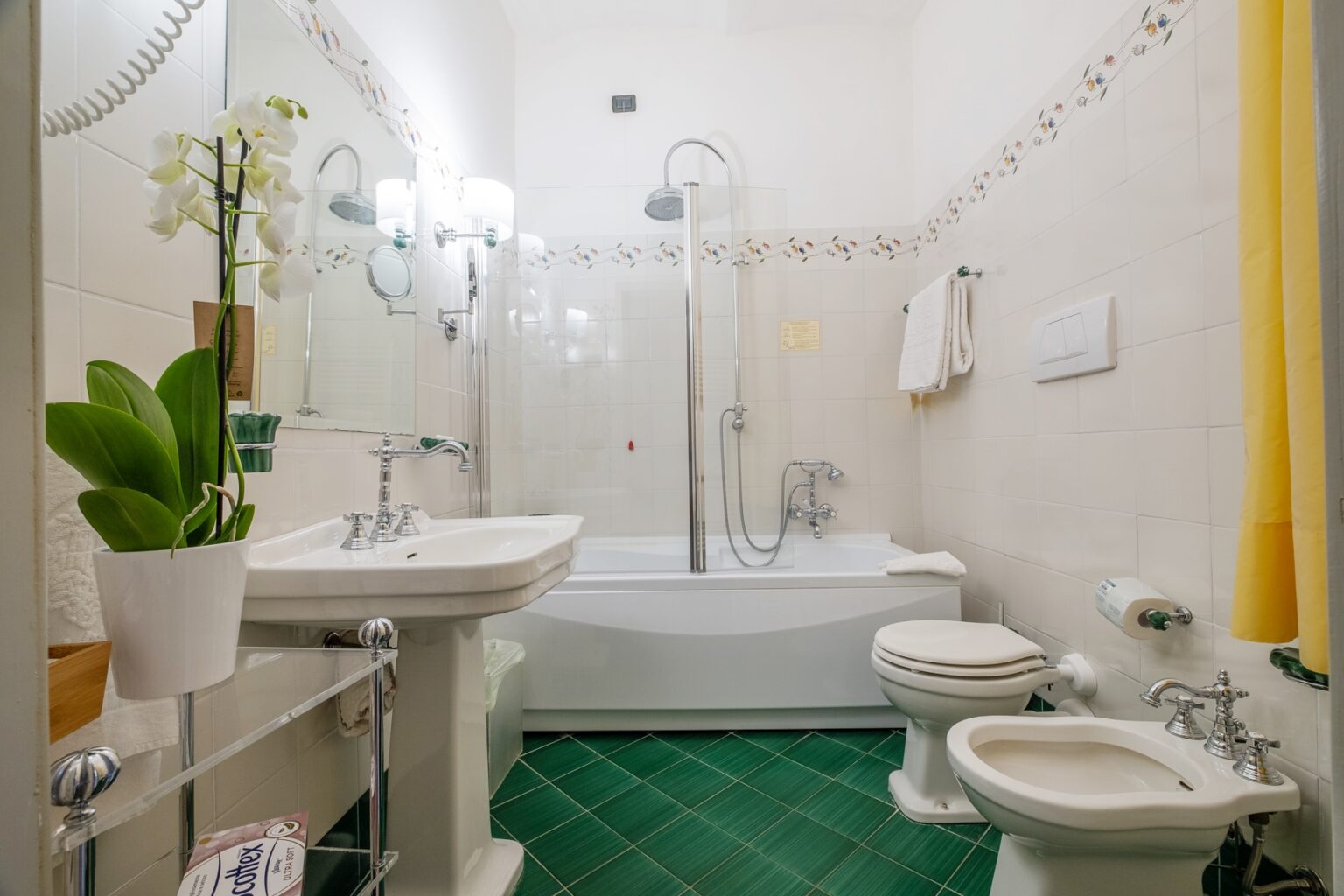 Modern bathroom featuring a white sink, elegant fixtures, a bathtub with a shower, and green tiled flooring. Decorated with a potted orchid and towels, creating a clean and inviting atmosphere. | Only on SopranoVillas