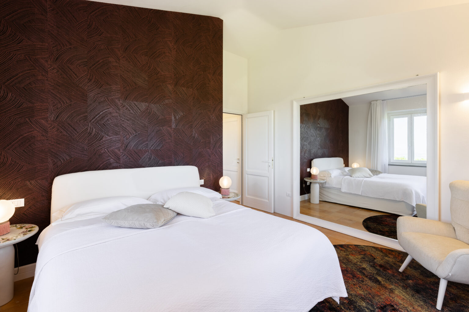 Modern bedroom featuring a white bed with decorative pillows, a textured dark accent wall, and a large mirror reflecting a cozy seating area. The room is well-lit with natural light and has stylish decor elements, creating a serene and inviting atmosphere. | Only on SopranoVillas