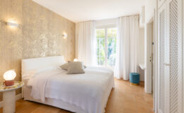 Bright and airy bedroom featuring a white bed with decorative pillows, a stylish side table with a lamp, and large windows draped in sheer curtains. The wall is adorned with elegant leaf-patterned wallpaper, enhancing the modern aesthetic of the space. A cozy blue stool and a white cabinet complete the room's design.