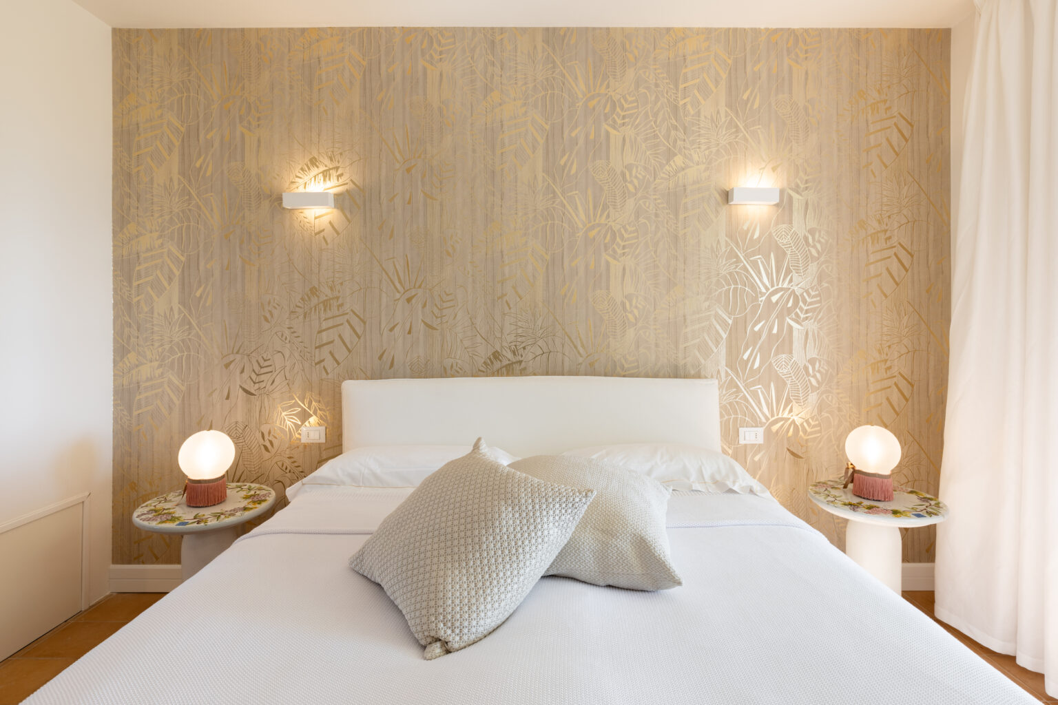 Modern bedroom interior featuring a white bed with decorative pillows, stylish side tables with unique lamps, and a gold leaf patterned wall, creating a serene and elegant atmosphere. | Only on SopranoVillas
