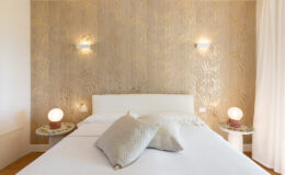 Modern bedroom interior featuring a white bed with decorative pillows, stylish side tables with unique lamps, and a gold leaf patterned wall, creating a serene and elegant atmosphere.