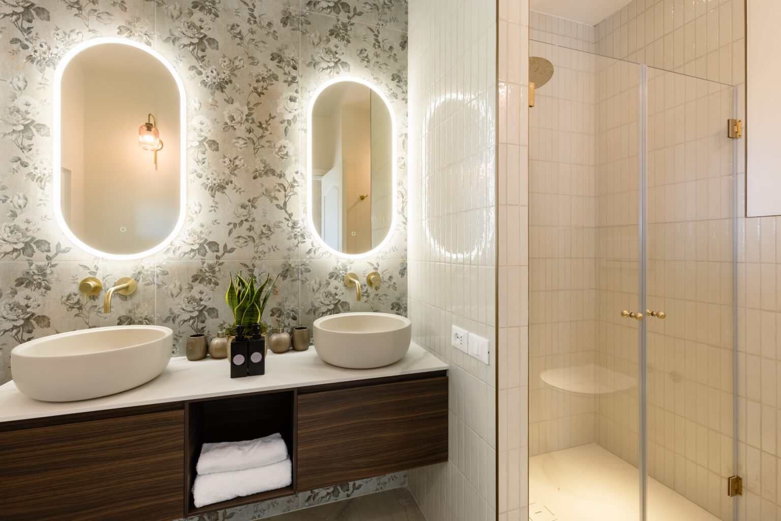 Modern bathroom featuring dual oval mirrors with LED lighting, stylish vessel sinks, and a glass shower enclosure. The decor includes floral wallpaper and elegant gold fixtures, complemented by a wooden vanity with neatly arranged towels and decorative plants. | Only on SopranoVillas