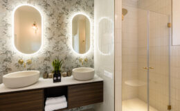 Modern bathroom featuring dual oval mirrors with LED lighting, stylish vessel sinks, and a glass shower enclosure. The decor includes floral wallpaper and elegant gold fixtures, complemented by a wooden vanity with neatly arranged towels and decorative plants.