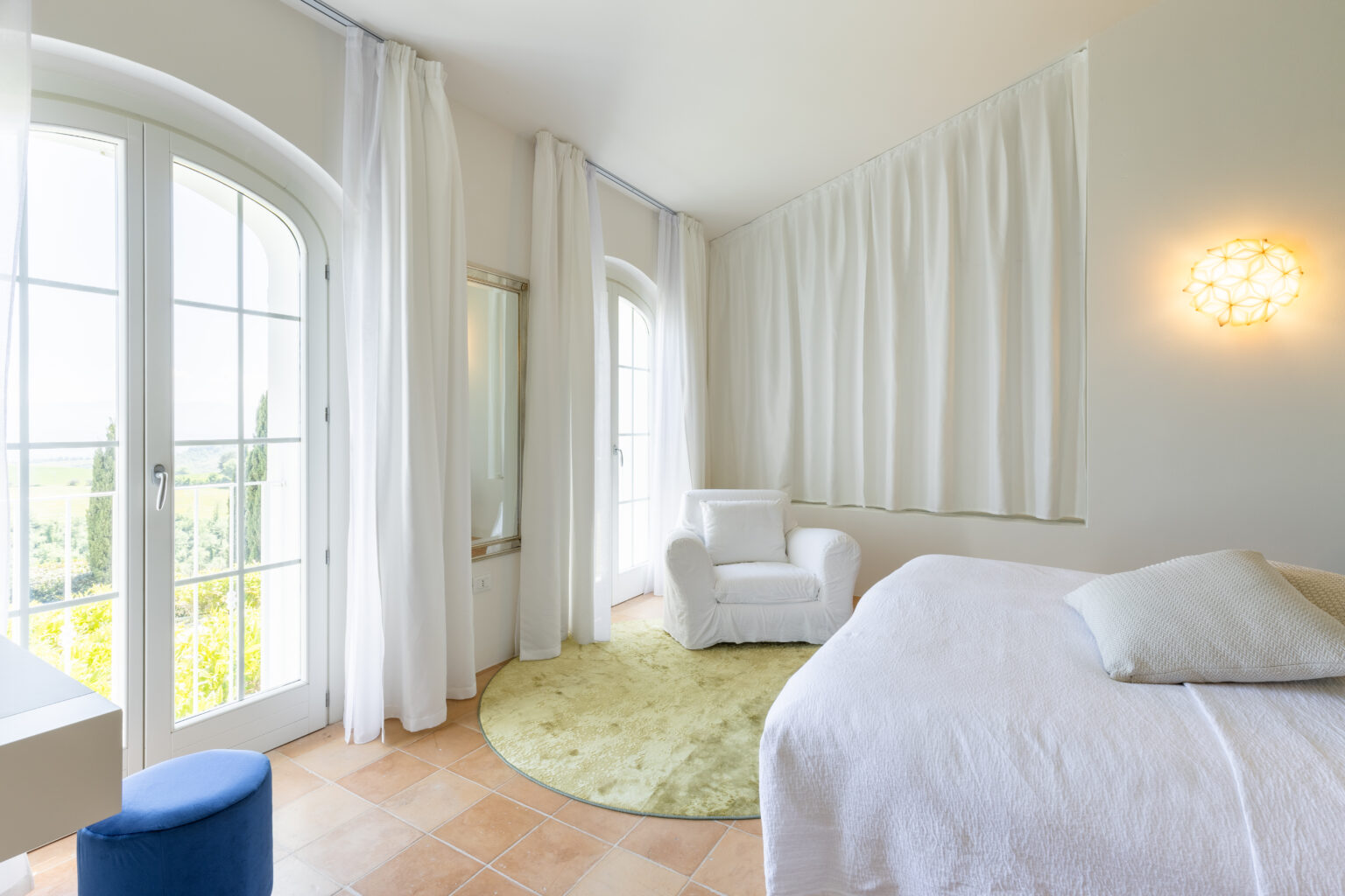 Bright and airy bedroom featuring large windows with white curtains, a cozy white armchair, and a round green rug. The room is decorated in neutral tones, creating a serene atmosphere perfect for relaxation. | Only on SopranoVillas