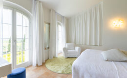 Bright and airy bedroom featuring large windows with white curtains, a cozy white armchair, and a round green rug. The room is decorated in neutral tones, creating a serene atmosphere perfect for relaxation.