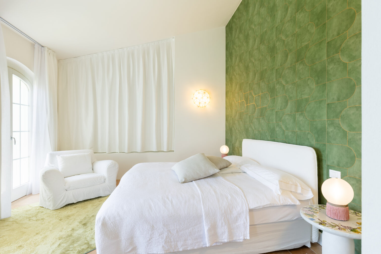 Modern bedroom interior featuring a white bed with soft linens, a cozy white armchair, and a vibrant green accent wall with unique tile design. Natural light filters through sheer curtains, enhancing the serene atmosphere. Decorative lighting adds a warm touch to the space. | Only on SopranoVillas