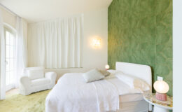 Modern bedroom interior featuring a white bed with soft linens, a cozy white armchair, and a vibrant green accent wall with unique tile design. Natural light filters through sheer curtains, enhancing the serene atmosphere. Decorative lighting adds a warm touch to the space.