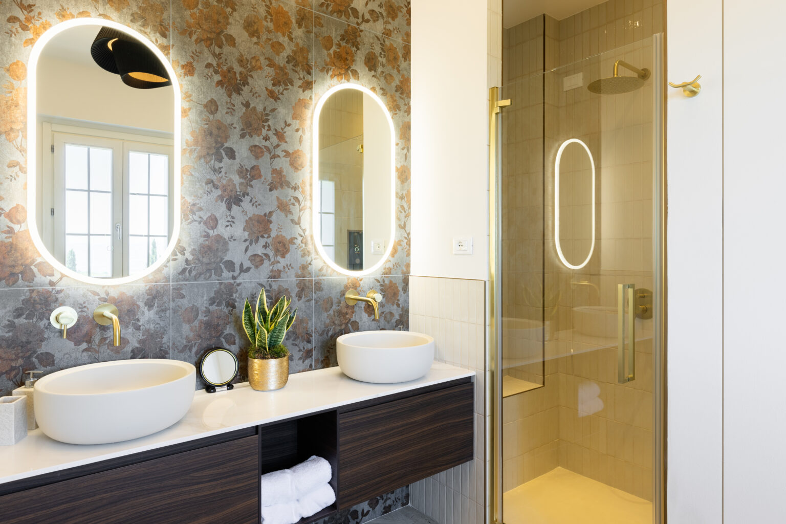 Modern bathroom design featuring dual oval mirrors with backlighting, stylish gold fixtures, white vessel sinks, and a glass shower enclosure. The wall is adorned with floral tiles, complemented by a green plant and neatly arranged towels in a wooden vanity. | Only on SopranoVillas