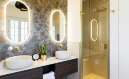 Modern bathroom design featuring dual oval mirrors with backlighting, stylish gold fixtures, white vessel sinks, and a glass shower enclosure. The wall is adorned with floral tiles, complemented by a green plant and neatly arranged towels in a wooden vanity.