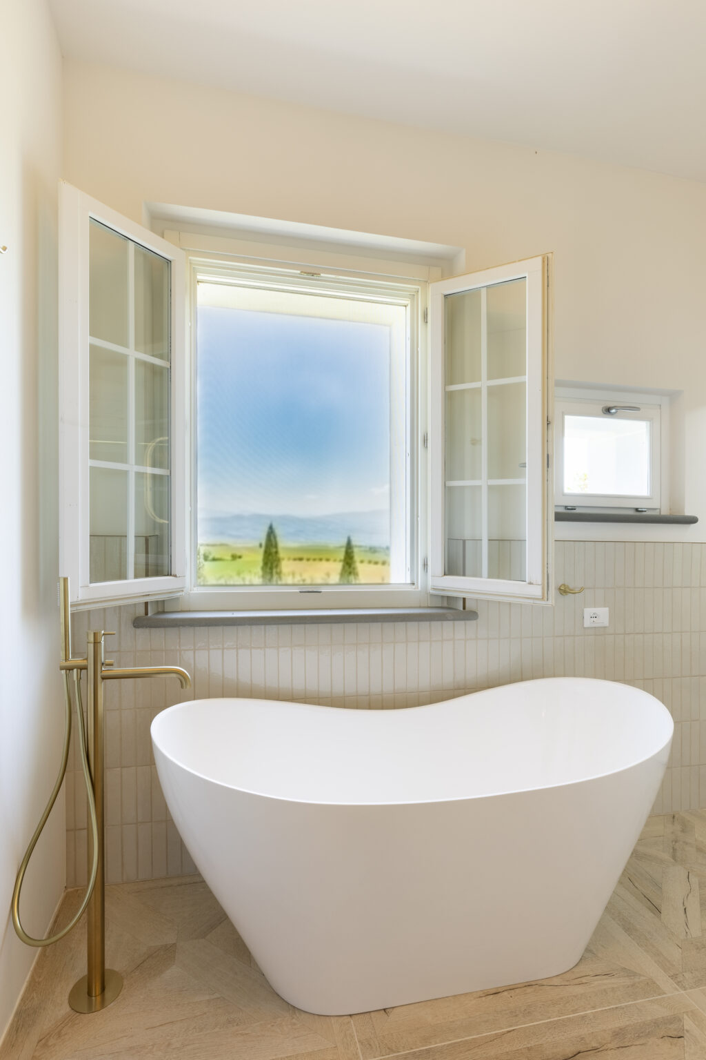 Modern bathroom featuring a freestanding white bathtub, elegant gold faucet, and large open windows with scenic countryside views. The interior showcases neutral tones and textured tile flooring, enhancing the serene atmosphere. | Only on SopranoVillas