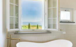 Modern bathroom featuring a freestanding white bathtub, elegant gold faucet, and large open windows with scenic countryside views. The interior showcases neutral tones and textured tile flooring, enhancing the serene atmosphere.