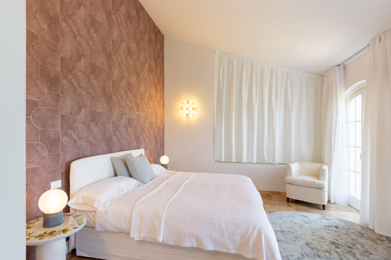 Modern bedroom featuring a white bed with soft linens, a textured pink accent wall, stylish lighting fixtures, and large windows draped in sheer curtains, creating a serene and inviting atmosphere. | Only on SopranoVillas