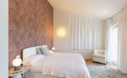 Modern bedroom featuring a white bed with soft linens, a textured pink accent wall, stylish lighting fixtures, and large windows draped in sheer curtains, creating a serene and inviting atmosphere.