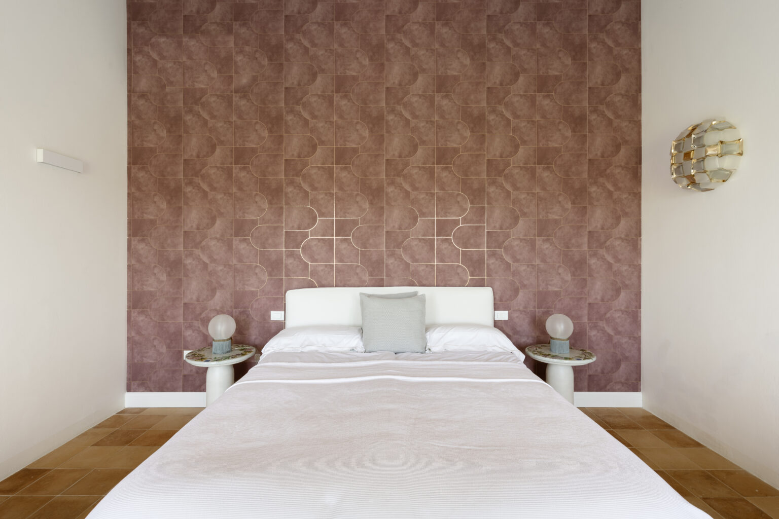 Modern bedroom interior featuring a white bed with a gray accent pillow, set against a textured mauve wall adorned with geometric patterns. Two stylish side tables with round lamps complement the decor, while warm-toned flooring adds to the cozy atmosphere. Ideal for home design inspiration. | Only on SopranoVillas