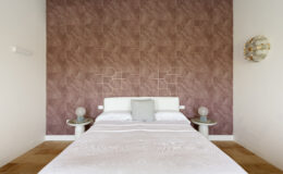 Modern bedroom interior featuring a white bed with a gray accent pillow, set against a textured mauve wall adorned with geometric patterns. Two stylish side tables with round lamps complement the decor, while warm-toned flooring adds to the cozy atmosphere. Ideal for home design inspiration.